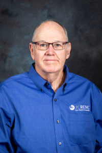 Headshot of a bald Caucasian man wearing eyeglasses. He is wearing a blue shirt with a JCREMC logo.