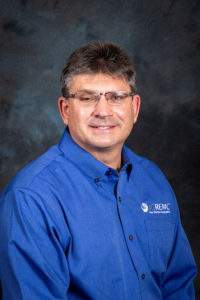 Headshot of a man with dark hair wearing glasses. He is wearing a blue dress shirt with a white JCREMC logo.