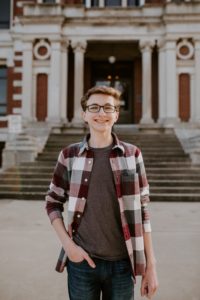 Andrew Hodges, a male high school senior with brown hair and glasses, stands in front of the Johnson County Courthouse wearing jeans and a plaid flannel shirt.