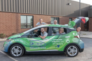 Five JCREMC employees in gray polo shirts sit in a Chevrolet Bolt electric car with a green and blue swirled wrap.