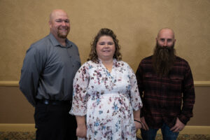 A tall bald man in a gray dress shirt, a short brunette woman in a white dress, and a short bald man with beard in a plaid shirt stand next to each other.