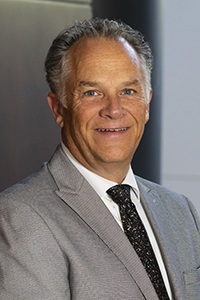 Headshot of a man with gray hair wearing a gray suit jacket, white dress shirt, and dark tie.