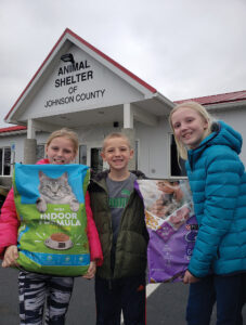 A young girl with blonde hair wears a pink coat and holds a large blue and green bag of cat food. A young lady with blonde hair wears a blue goat and holds a purple bag of pet food. A young boy with sandy hair stands between the girls wearing a black coat.
